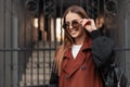 Happy stylish young woman with positive smile in fashion trench coat in trendy sunglasses with leather backpack poses near iron Royalty Free Stock Photo