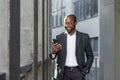 Happy successful African American businessman boss walking outside office building, manager holding smartphone reading Royalty Free Stock Photo