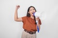 Happy successful Asian woman wearing a brown shirt raising clenched fist while shouting and screaming loud using megaphone speaker Royalty Free Stock Photo