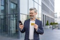 Happy and successful man in business suit outside office building, senior mature boss holding bank credit card and Royalty Free Stock Photo