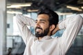 Happy and successful young Indian man, businessman and office worker sitting at workplace with hands behind head and Royalty Free Stock Photo