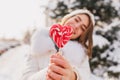 Happy sunny winter morning of joyful girl holding closeup pink heart lollypop on street. Sweet time, delicious, cold