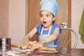 5 year old surprised girl in blue dress and dressed as chef in kitchen laughs and rolling out dough Royalty Free Stock Photo