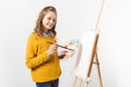 happy teen female painter in yellow sweater with paint brush and palette near easel with blank canvas