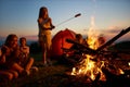 Happy teen girl frying sausage over fire, having fun with friends at campsite. Royalty Free Stock Photo