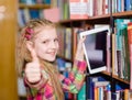 Happy teen girl pulls a tablet pc off the shelves in the library