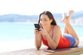 Happy teen using phone lying on a bench on the beach