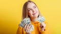 Happy teenage blonde girl holding in hands money cash dollars dreaming thoughtfully isolated on color yellow background