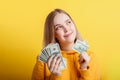 Happy teenage blonde girl holding in hands money cash dollars dreaming thoughtfully isolated on color yellow background. Portrait