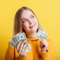 Happy teenage blonde girl holding in hands money cash dollars dreaming thoughtfully isolated on color yellow background