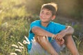 Happy teenage boy in blue t-shirt sits on the grass, dreaming, in backlight Royalty Free Stock Photo