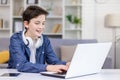 Happy teenage boy with headphones around his neck, sitting at a laptop in a bright room, tapping on a keyboard, playing Royalty Free Stock Photo