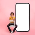 Happy thinking young mixed race lady with curly hair in casual sits on chair near huge smartphone with empty screen