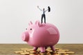 Happy tiny businessman celebrating success while standing on pink piggy bank and golden coins. White concrete wall background. Royalty Free Stock Photo