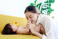 Happy toddle baby lying on yellow sofa in living room, mom kissing her little girl daughter at finger hand, parent playing with Royalty Free Stock Photo