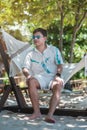 Happy traveler man with eyeglasses enjoy beautiful sea view, young man sitting on cradle at sand beach. Freedom, relaxing, Royalty Free Stock Photo