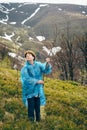 Happy traveller senior woman in blue rain jacket and jeans in mountains surrounded by forest with young plant in hands