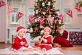 Happy twin babies dressed as Santa Claus are sitting near Christmas tree at home in living room Royalty Free Stock Photo