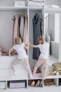 Family wardrobe. Happy twin sisters on shelve in closet with natural organic clothes in cozy dressing room play joyfully