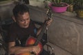 A happy and upbeat disabled asian man plays the guitar at the porch of his unfinished home. A paraplegic guitarist