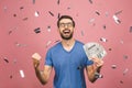 Happy winner! Young rich man in casual t-shirt holding money dollar bills with surprise isolated over pink background