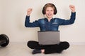 Happy winning teenager boy working or playing on laptop computer while sitting on the floor with legs crossed with headphones on Royalty Free Stock Photo