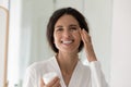 Happy woman in bathrobe holds jar applying cream on cheek Royalty Free Stock Photo