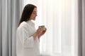 Happy woman with cup of drink near window at home, space for text. Lazy morning Royalty Free Stock Photo