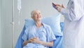 Happy woman doctor talking and using tablet to sick old female senior elderly patient lying in bed in hospital ward room in Royalty Free Stock Photo