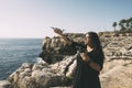 happy woman with drone, looking to the side black dress and remote control, on cliff, by the sea sunny day. technological concept