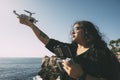 happy woman with drone, looking to the side black dress and remote control, on cliff, by the sea sunny day. technological concept