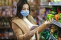 Happy woman in face mask hold phone and choose vegetables in supermarket. Beautiful young business woman choosing