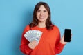 Happy woman with fan of banknotes and phone in hands looking directly at camera with satisfied expression, dark haired lady wins