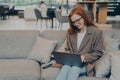 Woman in glasses sitting on cozy couch in office lounge while chatting on computer laptop