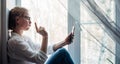 Happy woman in glasses and white shirt sits on the windowsill, holding smartphone in her hand. Cute blonde smiles at phone camera Royalty Free Stock Photo