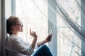 Happy woman in glasses and white shirt sits on the windowsill, holding smartphone in her hand. Cute blonde smiles at phone camera Royalty Free Stock Photo