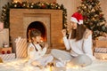 Happy woman with her little daughter sitting in living room near christmas tree and fireplace with gift boxes, being very glad, Royalty Free Stock Photo