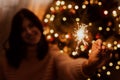 Happy woman holding firework at christmas tree with golden lights. Stylish girl with burning sparkler celebrating in festive dark Royalty Free Stock Photo