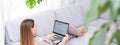 Young or middle age woman sitting with laptop on grey couch in home office with monstera plant. Concept of remote Royalty Free Stock Photo