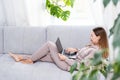 Young or middle age woman sitting with laptop on grey couch in home office with monstera plant. Concept of remote Royalty Free Stock Photo