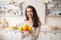 Happy woman holding plate with fresh fruits, looking and smiling at camera, standing in kitchen interior Royalty Free Stock Photo