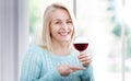 Happy woman holds a glass of red wine and looks relaxed at the camera sitting near the window. Royalty Free Stock Photo