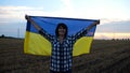 Happy woman looks into camera standing on wheat field with a lifted blue-yellow banner at sunset. Ukrainian smiling lady