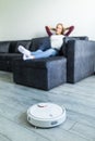 People, housework and technology concept. Happy woman lying on the couch while robot vacuum cleaner clean home Royalty Free Stock Photo
