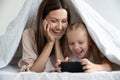 Happy woman lying under blanket with daughter, playing mobile games. Royalty Free Stock Photo