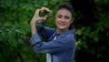 Happy woman making heart sign by hands in front of tree leaves, care of nature