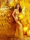 Happy Woman in Party Golden Dress. Sexy Girl in Sequin Long Gown over Glitter Gold Wall Background with Yellow Star Balloon. Royalty Free Stock Photo