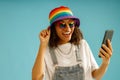 Happy woman in rainbow cap is holding mobile phone and looking at camera over blue studio background Royalty Free Stock Photo