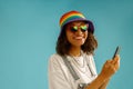 Happy woman in rainbow cap is holding mobile phone and looking at camera over blue studio background Royalty Free Stock Photo