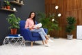 Happy woman reading book in armchair. Indoor plants for trendy interior design